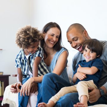 Happy multiethnic family sitting on sofa laughing together. Cheerful parents playing with their sons at home. Black father tickles his little boy while the mother and the brother smile.